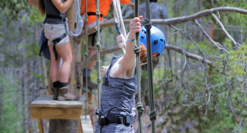 A person wearing safety gear is secured by ropes as they navigate an obstacle in a ropes course. 